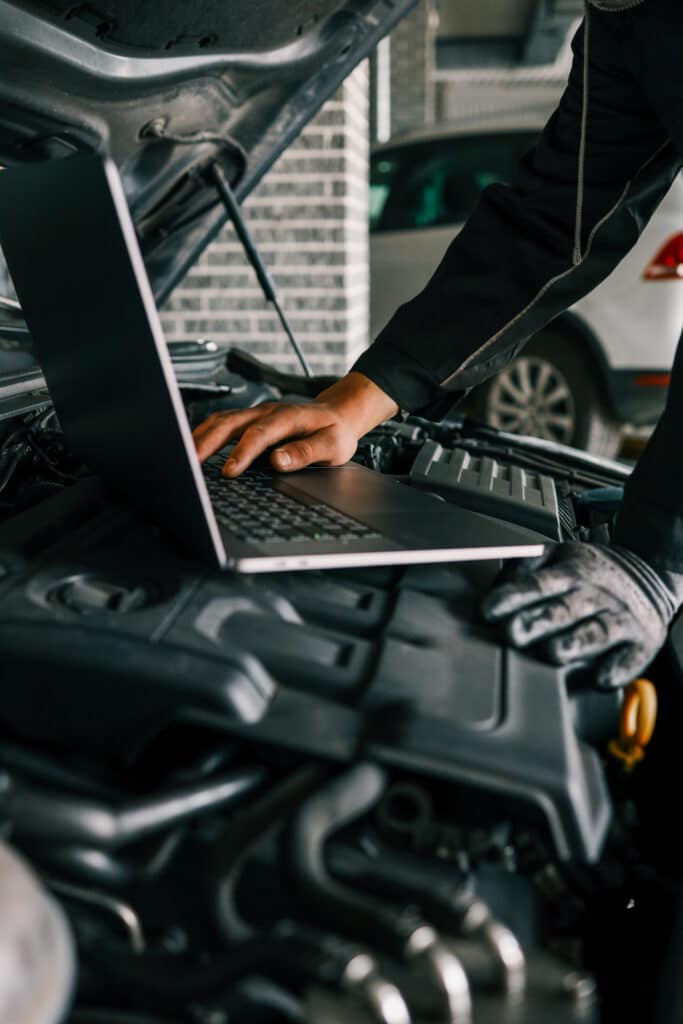 Mécanicien avec gant saisissant un ordinateur portable posé sur le moteur d'une voiture ouverte, effectuant un diagnostic électronique.