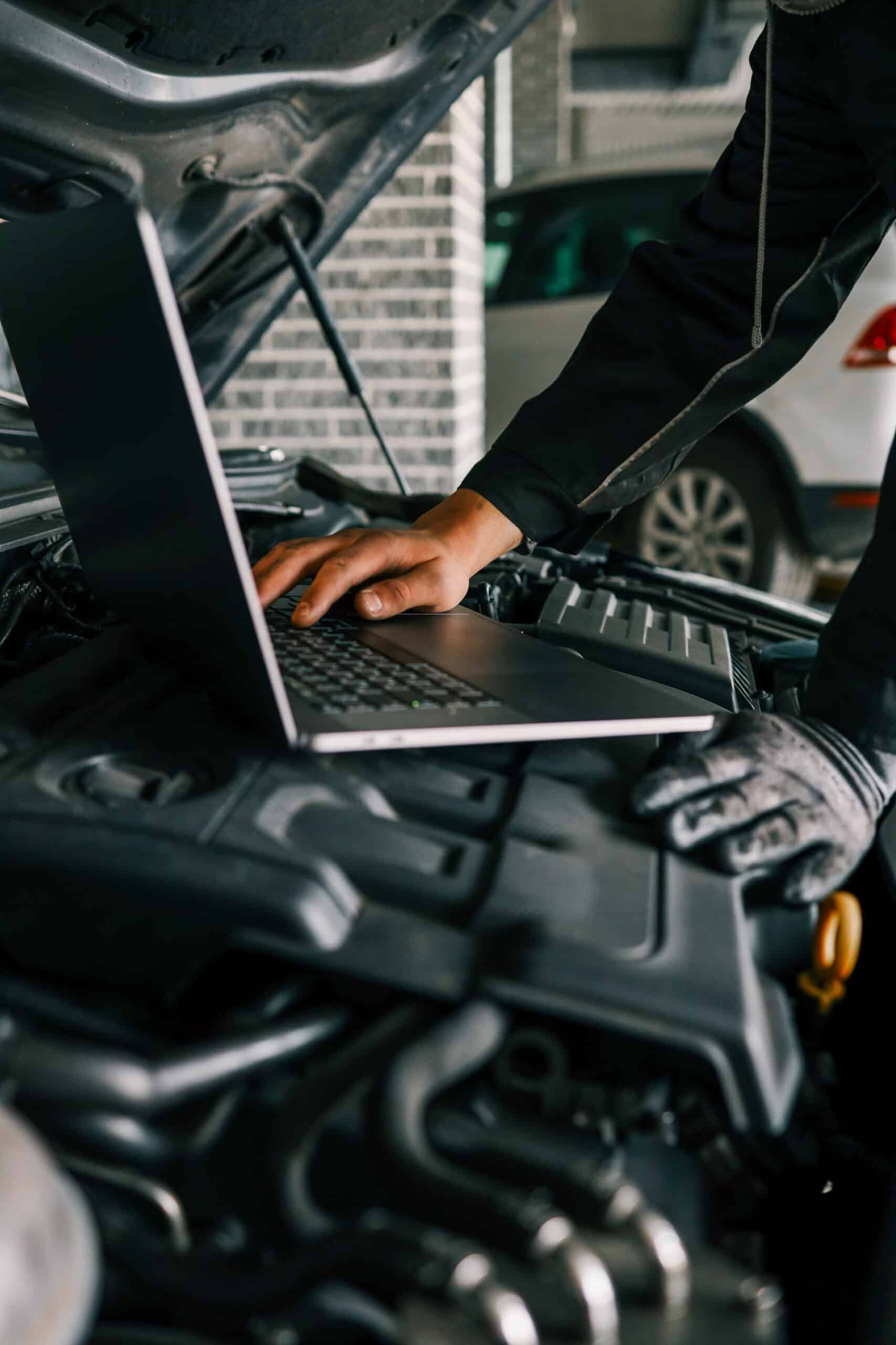 Mécanicien avec gant saisissant un ordinateur portable posé sur le moteur d'une voiture ouverte, effectuant un diagnostic électronique.