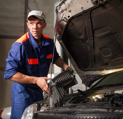 Jeune mécanicien en combinaison bleue avec des bandes orange inspectant le moteur d'une voiture, tenant une pièce noire sous le capot ouvert.