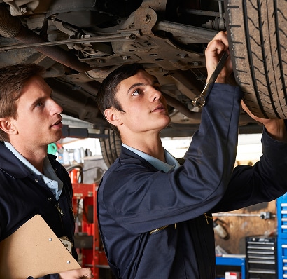 Deux mécaniciens en uniforme bleu inspectent la voiture soulevée, l'un utilisant une clé sur un pneu, l'autre tenant un presse-papiers.