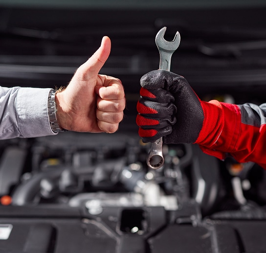 Mécanicien et Client Satisfaits : Réparation Auto Main d'un client pouce levé à côté de la main gantée d'un mécanicien tenant une clé, devant un moteur de voiture ouvert.
